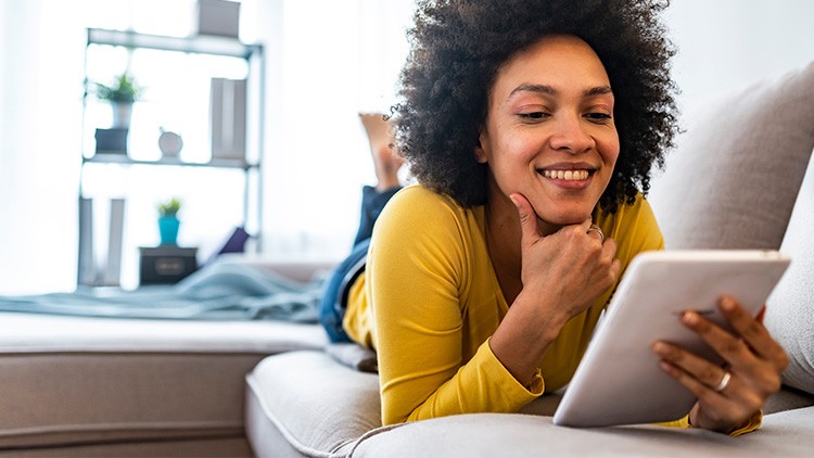 Person in gelbem Oberteil liegt auf einem Sofa und hält ein Tablet, im Hintergrund steht ein Regal mit Pflanzen und Deko.
