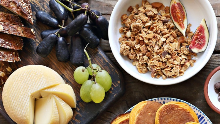 Ein Holztisch mit Granola, frischen Feigen, Brot, Käse, Trauben, Tomaten, Pancakes, einer kleinen Kürbisfrucht und Besteck.