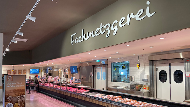 Eine Fleischtheke mit verschiedenen Fleisch- und Wurstwaren, dahinter Mitarbeitende in weißer Kleidung, links ein Info-Aufsteller.