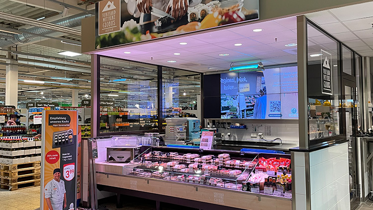 Frischetheke mit verpackten Fleischprodukten, Werbesäule mit Angebot und Schriftzug GLOBUS, Küchenausstattung im Hintergrund.