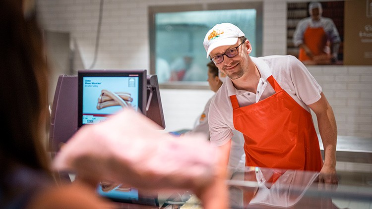 Ein Mitarbeitender mit orangefarbener Schürze reicht hinter einer Theke ein Produkt, im Hintergrund ist ein Bildschirm mit Würstchen zu sehen.