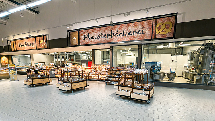Meisterbäckerei und Konditorei mit Regalen voller Backwaren, Glasfront mit Blick in die Backstube, Schriftzug auf Backsteinwand.