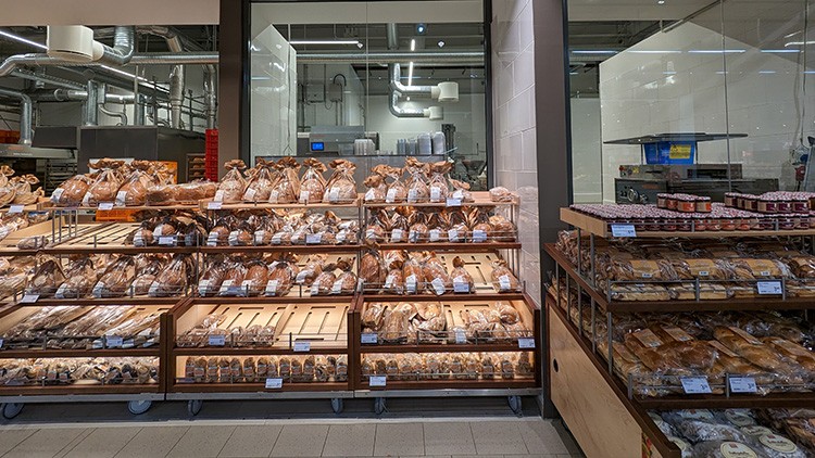 Regale mit verschiedenen Brotsorten, teils in Tüten verpackt, vor einer gläsernen Backstube mit sichtbaren Maschinen.
