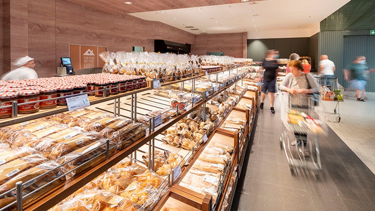 Regale mit Brot und Backwaren in einer GLOBUS-Filiale, daneben Kunden mit Einkaufswagen in einem breiten Gang.