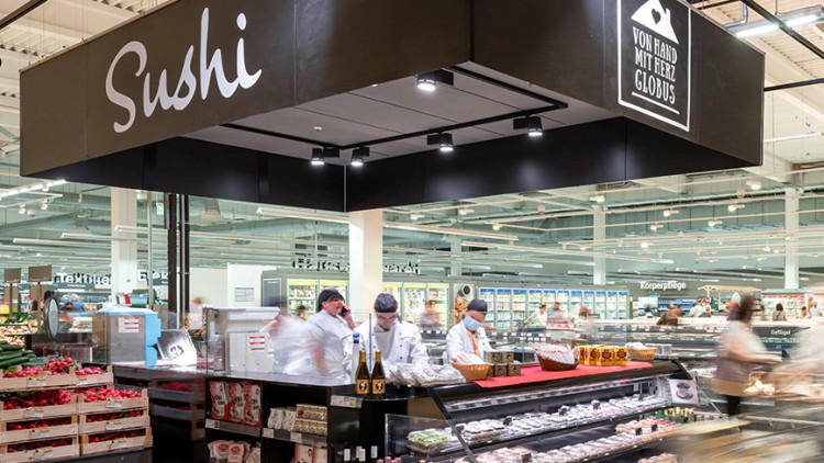 Eine Sushi-Theke mit frischen Produkten und Mitarbeitern, darüber ein Schild mit der Aufschrift „Von Hand mit Herz GLOBUS“.