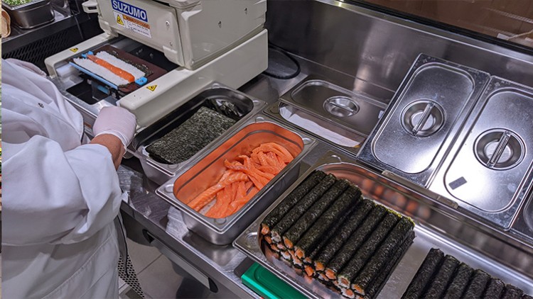 Ein Sushi-Zubereitungsbereich mit Nori-Blättern, Lachsstreifen, fertigen Maki-Rollen und einer SUZUMO-Maschine.