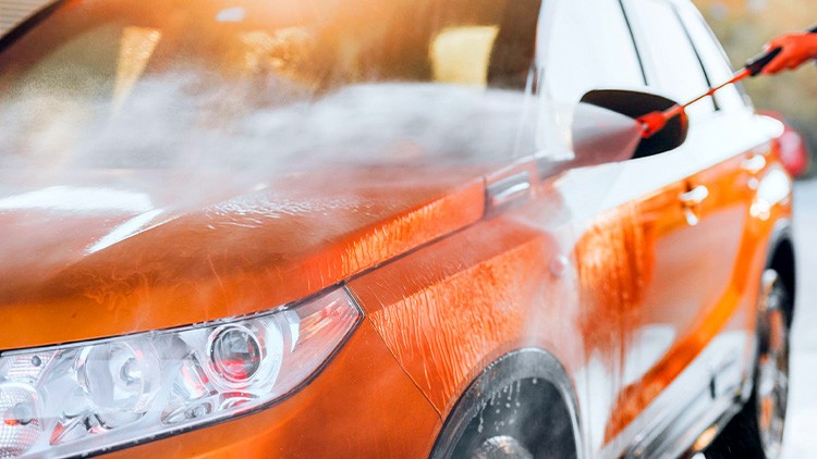 Ein orangefarbenes Auto wird mit einem Hochdruckreiniger abgespritzt, Wasser und Schaum bedecken den Lack.