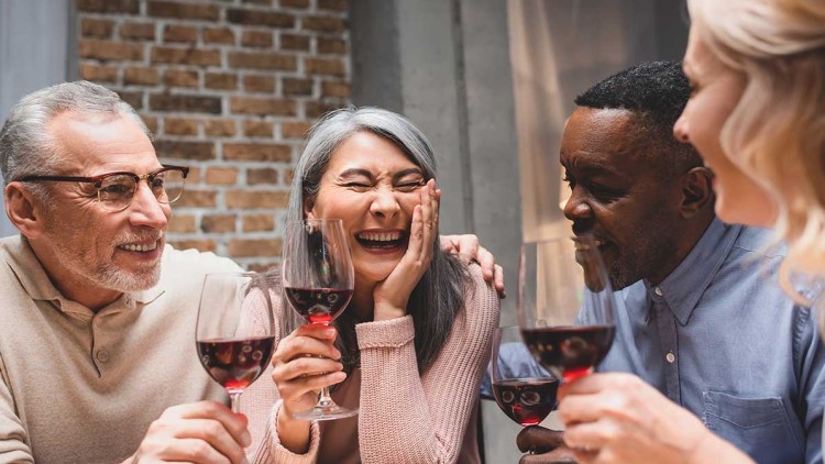 Vier Personen sitzen an einem Tisch mit Gläsern Rotwein und einer Backsteinwand im Hintergrund.