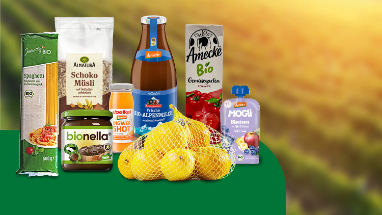 Verschiedene Bio-Lebensmittel, darunter Spaghetti, Müsli, Saft, Brotaufstrich, Äpfel, Blaubeerpüree und Tomatensaft vor einem Feld.