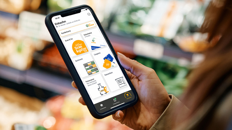 Eine Hand hält ein Smartphone mit geöffneter GLOBUS-App, im Hintergrund sind Regale mit unscharfem Gemüse zu sehen.