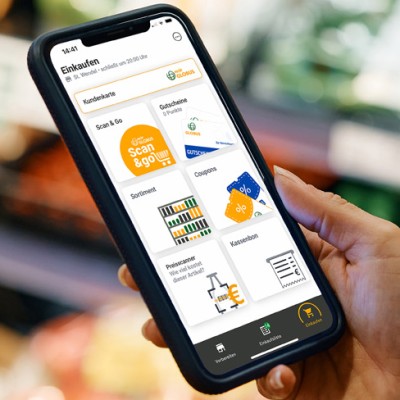 Eine Hand hält ein Smartphone mit geöffneter GLOBUS-App, im Hintergrund sind Regale mit unscharfem Gemüse zu sehen.