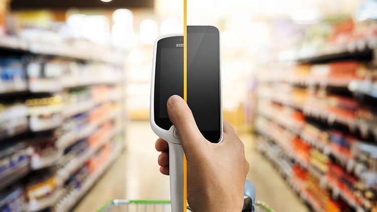 Eine Hand hält ein Scan & Go-Gerät, im Hintergrund sind unscharfe Supermarktregale mit Waren zu sehen.