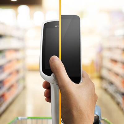 Eine Hand hält ein Scan & Go-Gerät, im Hintergrund sind unscharfe Supermarktregale mit Waren zu sehen.