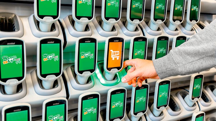 Eine Hand entnimmt ein Scan & Go Gerät mit orangefarbenem Einkaufswagensymbol aus einer Ladestation mit weiteren grünen Displays.