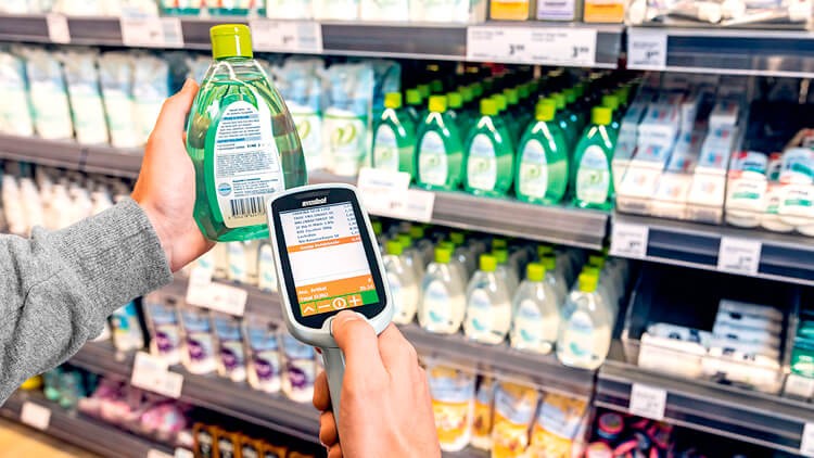 Eine Hand hält ein Scan & Go-Gerät, das ein Produkt scannt, während eine andere Hand eine grüne Flasche vor einem Regal hält.