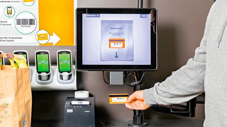 Eine Person scannt die mein GLOBUS Karte an einem Selbstbezahlautomaten mit Bildschirm, Scanner und Einkaufstasche mit Obst.