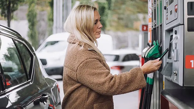 Person an einer Tankstation bedient mit einer Karte die Zapfsäule, das Fahrzeug steht daneben, im Hintergrund geparkte Autos.