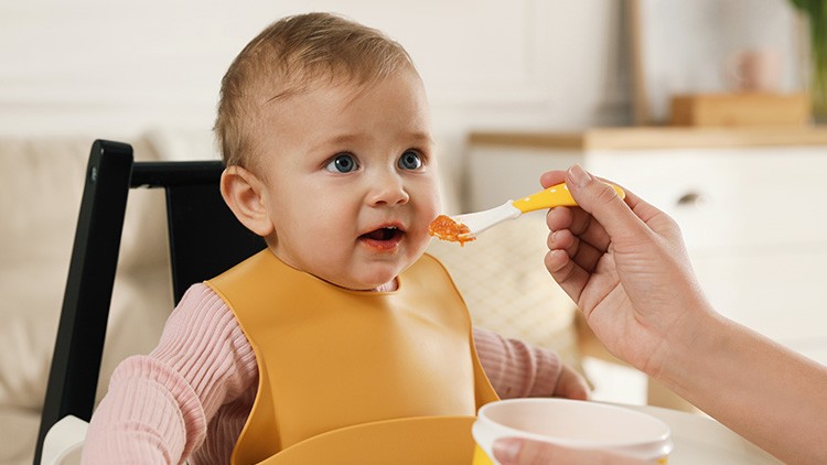 Eine Hand füttert ein Kind in einem Hochstuhl mit einem gelben Löffel, das ein gelbes Lätzchen trägt.