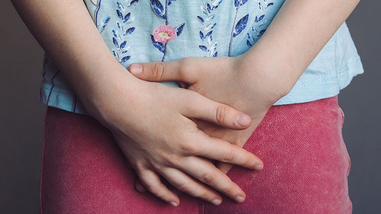 Eine Person in blauer Bluse mit Blumenmuster und rosa Hose hält die Hände gefaltet vor ihrem Körper.
