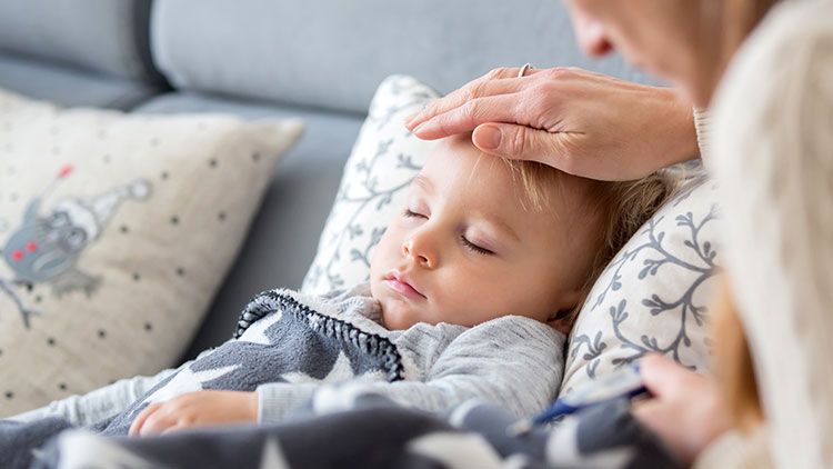Hand streicht beruhigend über ein Kind, das auf einem Sofa mit gemusterten Kissen und einer Decke liegt.