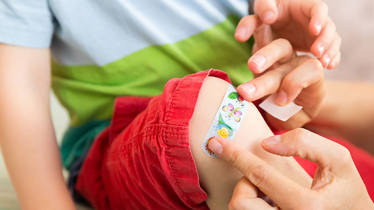 Eine Hand klebt ein bunt bedrucktes Pflaster mit Blumenmotiv auf das Knie eines Kindes in roten Shorts.