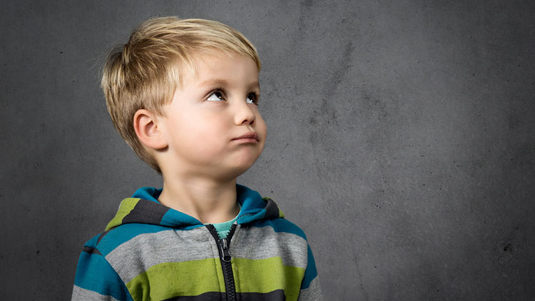 Kind mit blondem Haar trägt einen Kapuzenpullover in Blau, Grün und Grau vor einer grauen Betonwand.