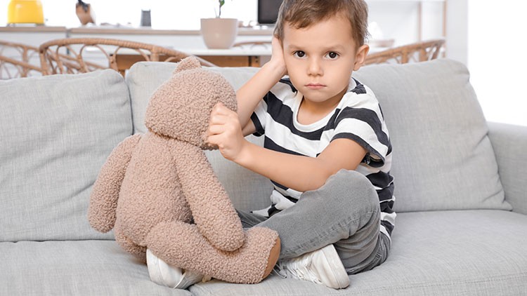 Kind sitzt auf einem grauen Sofa und hält einen braunen Plüschbären mit beiden Händen.