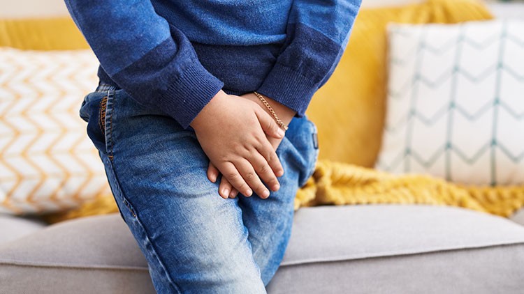 Kind in blauer Kleidung hält mit beiden Händen den Bauchbereich und steht vor einem Sofa mit gelbem Plaid und gemusterten Kissen.