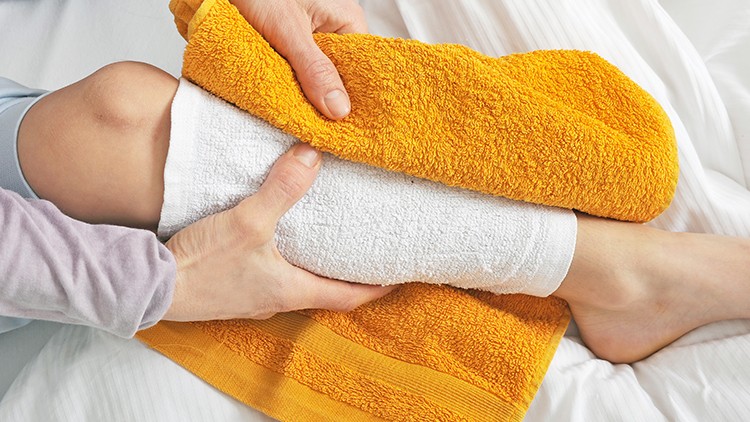 Eine Hand hält ein oranges Handtuch auf einem in ein weißes Handtuch gewickelten Bein, das auf einem Bett liegt.