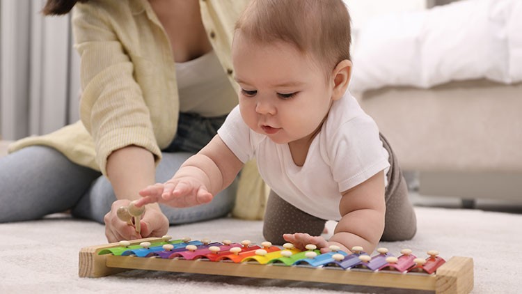Ein Baby in weißem Shirt spielt auf dem Boden mit einem bunten Xylophon aus Holz, daneben kniet eine erwachsene Person.