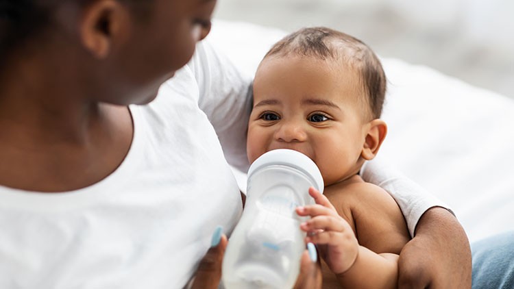 Eine Person hält ein Baby im Arm, das aus einer transparenten Flasche mit Milch trinkt.