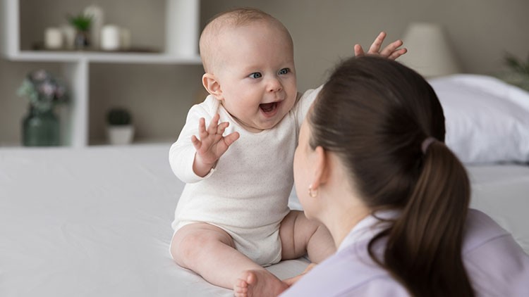 Ein Baby im weißen Strampler sitzt auf einem Bett und streckt die Arme nach einer Person mit braunen Haaren aus.