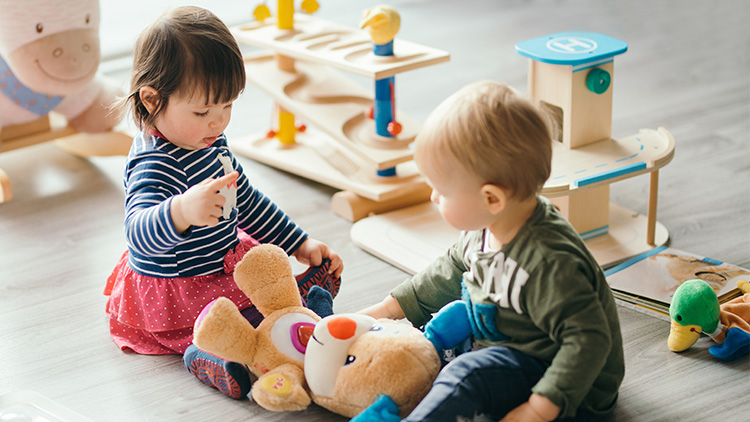Kinder spielen auf einem Holzboden mit Plüschtieren, Holzspielzeug und einem rosa Kunststoffkorb im Hintergrund.