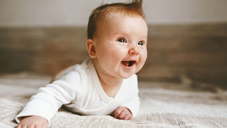 Ein Baby in weißem Oberteil liegt auf einem Bett mit Holzkopfteil im Hintergrund.