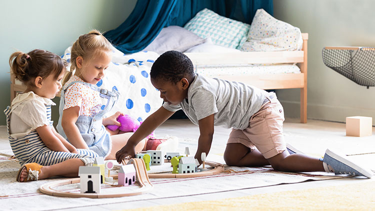 Drei Kinder spielen auf einem hellen Teppich mit kleinen Holzspielzeugen in einem bunt dekorierten Kinderzimmer.