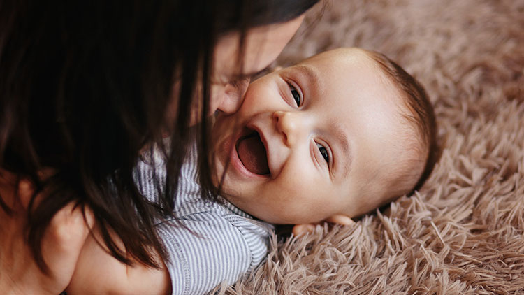 Person mit dunklen Haaren hält ein Baby in gestreiftem Outfit auf einem beigen, flauschigen Teppich.