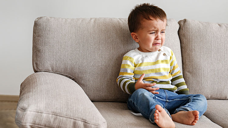 Kind in gestreiftem Pullover und blauer Jeans sitzt auf einem beigen Sofa vor einer hellen Wand.