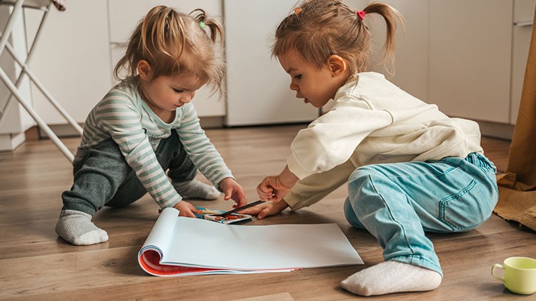 Zwei Kinder sitzen auf einem Holzboden und betrachten gemeinsam ein aufgeschlagenes großes Buch, daneben steht eine gelbe Tasse.