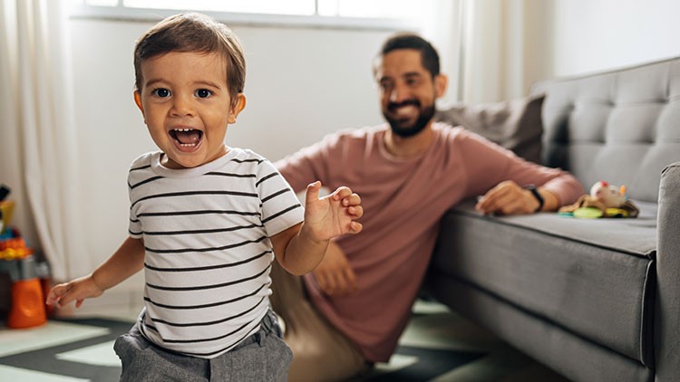 Ein Kind in gestreiftem Oberteil läuft auf einem Teppich, im Hintergrund sitzt eine Person neben einem grauen Sofa.