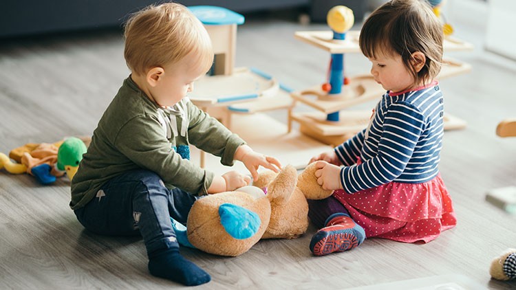 Zwei Kleinkinder sitzen auf einem Holzboden und spielen mit einem braunen Plüschbären in einer Umgebung mit Holzspielzeug.