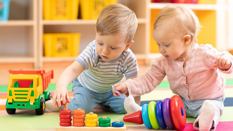 Zwei Kinder spielen auf einer bunten Matte mit einem Spielzeug-LKW, bunten Stapelringen und farbigen Steckringen.