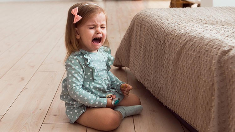 Kind in gepunktetem Kleid und Socken sitzt auf einem Holzboden neben einem Bett mit strukturierter Decke.