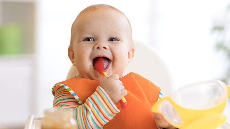 Ein Kind in einem Hochstuhl trägt ein orangefarbenes Lätzchen und hält einen gelben Löffel vor einer verschwommenen Pflanze.