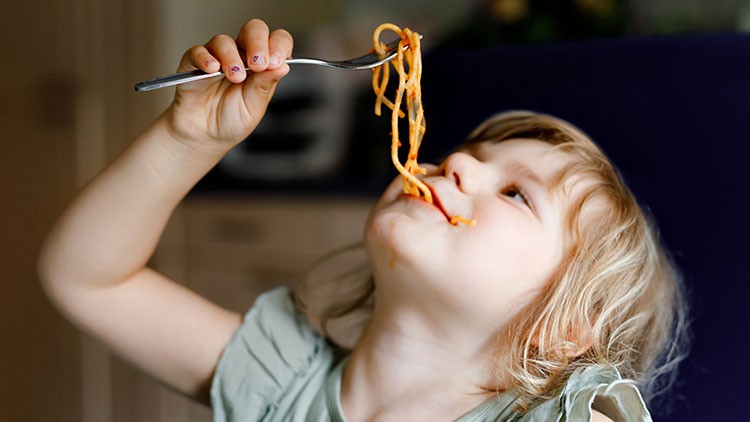Eine Kinderhand hält eine Gabel mit Spaghetti, im Hintergrund ist eine unscharfe Küche erkennbar.