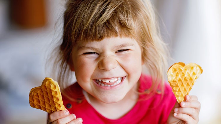 Kind in rotem Oberteil hält zwei herzförmige Waffeln in den Händen, Hintergrund unscharf.