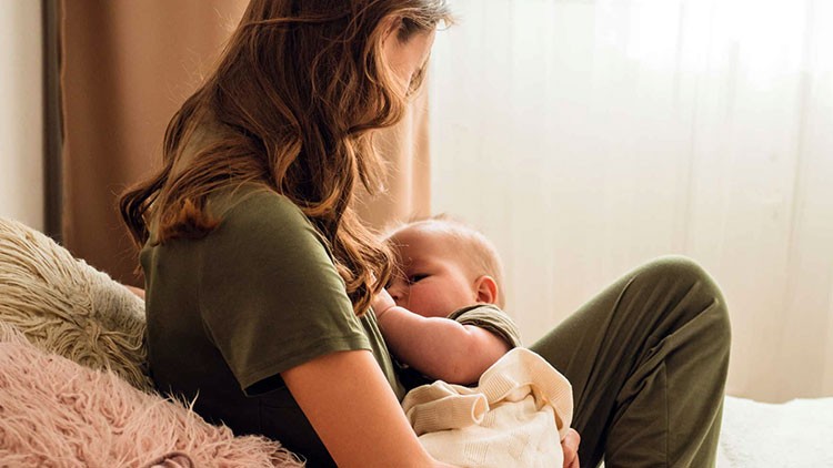 Person in grüner Kleidung sitzt auf einem Sofa mit Kissen und hält ein Baby in einer cremefarbenen Decke im Arm.