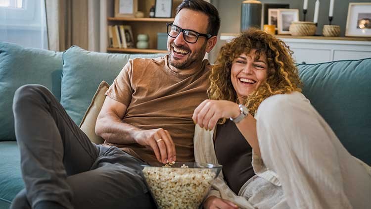 Zwei Personen sitzen auf einem Sofa mit einer Schüssel Popcorn, im Hintergrund ein Regal mit Deko und Kerzen.