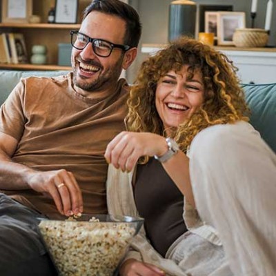 Zwei Personen sitzen auf einem Sofa mit einer Schüssel Popcorn, im Hintergrund ein Regal mit Deko und Kerzen.