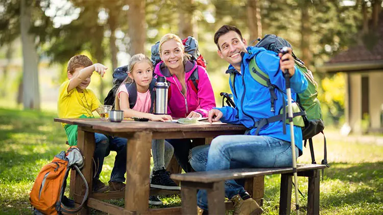 Vier Personen sitzen mit Wanderrucksäcken an einem Holztisch im Park, eine hält eine Thermoskanne, Bäume stehen im Hintergrund.
