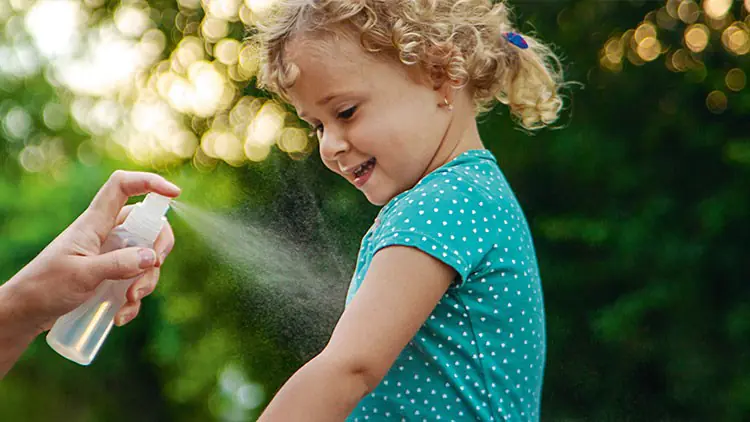 Eine Hand sprüht mit einer Sprühflasche Flüssigkeit auf den Arm eines Kindes in einem grünen Kurzarmshirt, Hintergrund: Natur.